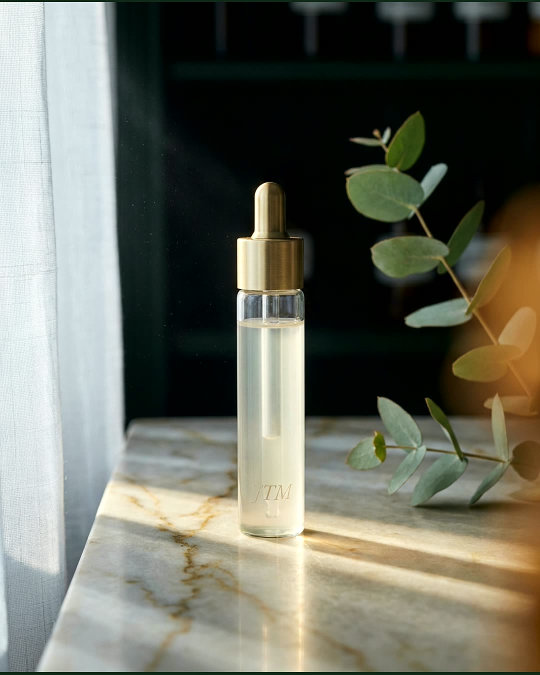 Editorial still life: a brushed-gold dropper vial on veined marble, with a soft eucalyptus drape — JTM Aesthetics.